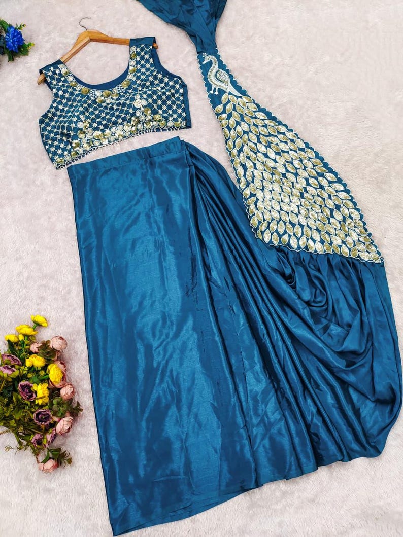 May include: A teal blue sleeveless blouse with a white lattice pattern and floral embroidery, paired with a matching teal blue skirt featuring a long, flowing panel decorated with a large white and gold peacock feather motif and sequins.