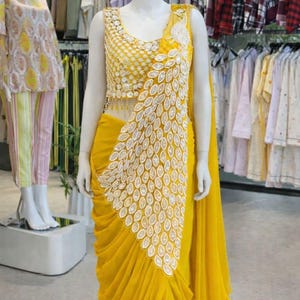 May include: A bright yellow sleeveless blouse and matching draped skirt ensemble. The bodice features intricate gold embroidery and beadwork. A large, white, leaf-shaped appliqu&eacute; pattern cascades diagonally across the front of the garment.