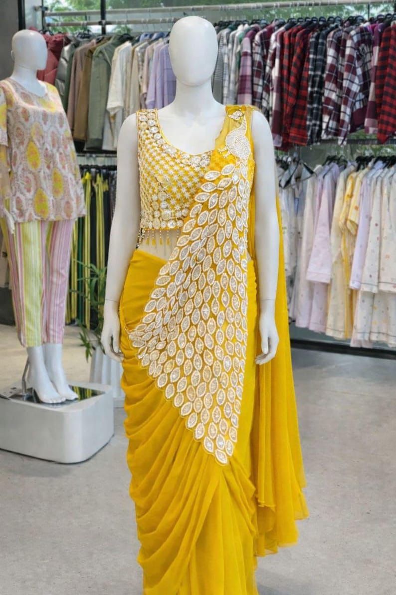 May include: A bright yellow sleeveless blouse and matching draped skirt ensemble. The bodice features intricate gold embroidery and beadwork. A large, white, leaf-shaped appliqu&eacute; pattern cascades diagonally across the front of the garment.