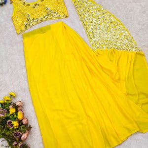 May include: A bright yellow sleeveless blouse and a matching long skirt. The blouse features intricate silver embroidery and mirror work. The skirt includes a long, flowing panel decorated with a silver peacock motif and leaf-shaped mirror embellishments.