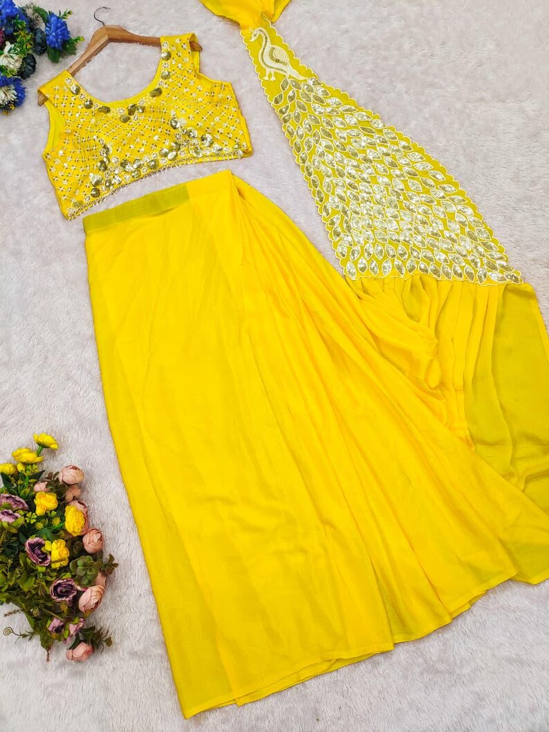 May include: A bright yellow sleeveless blouse and a matching long skirt. The blouse features intricate silver embroidery and mirror work. The skirt includes a long, flowing panel decorated with a silver peacock motif and leaf-shaped mirror embellishments.