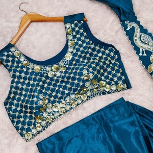 May include: A teal sleeveless blouse featuring a gold grid pattern and gold sequin embellishments, paired with a matching teal skirt. A separate teal fabric piece displays a white embroidered peacock design with scalloped edges.