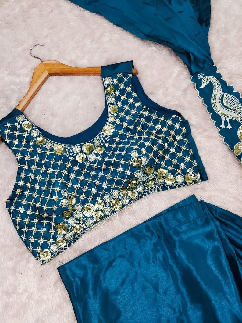 May include: A teal sleeveless blouse featuring a gold grid pattern and gold sequin embellishments, paired with a matching teal skirt. A separate teal fabric piece displays a white embroidered peacock design with scalloped edges.