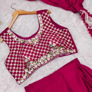 May include: A magenta sleeveless crop top featuring a white grid pattern and gold sequin embellishments, paired with a matching magenta skirt and a draped magenta fabric piece with a white embroidered peacock motif along the scalloped edge.