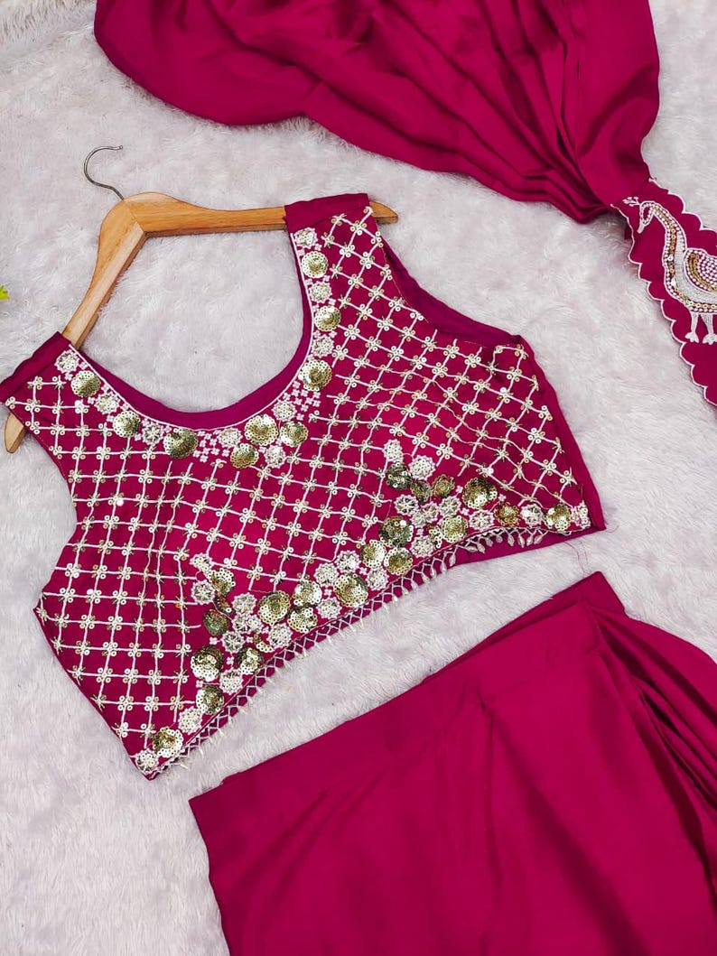 May include: A magenta sleeveless crop top featuring a white grid pattern and gold sequin embellishments, paired with a matching magenta skirt and a draped magenta fabric piece with a white embroidered peacock motif along the scalloped edge.