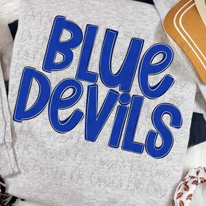 May include: A light gray long-sleeve shirt featuring the text Blue Devils in bold, royal blue letters with a dark blue outline. The shirt is displayed alongside a tan hat with white stripes and a white fabric with a brown football pattern.