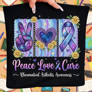 Op de afbeelding: Zwart kussen met een ontwerp met een vredesteken, hart en lint, allemaal met een galaxypatroon. De woorden "Peace Love Cure" en "Rheumatoid Arthritis Awareness" worden ook weergegeven.