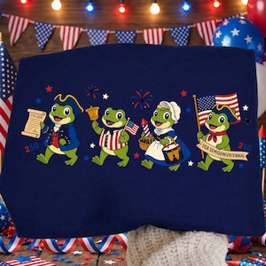 Könnte beinhalten: Marineblaues T-Shirt mit einem Design von vier Cartoon-Fröschen in patriotischer Kleidung. Jeder Frosch hält einen Gegenstand wie eine Schriftrolle, eine Glocke, eine Flagge und ein Banner mit der Aufschrift "USA Semiquincentennial" und der Zahl "250". Das T-Shirt ist mit Feuerwerk und Sternen verziert.
