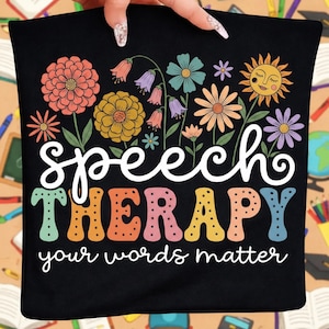 May include: Black tote bag featuring a floral design with the words "Speech Therapy" in white script and "THERAPY" in colorful block letters. The phrase "your words matter" is written below. The design includes a smiling sun and various flowers.