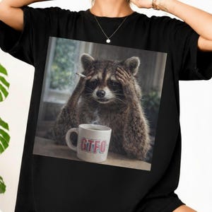 Può includere: T-shirt nero con un'immagine umoristica di un procione che fuma una sigaretta e si tiene la testa. Una tazza bianca con le lettere "GTFO" è di fronte al procione. L'immagine ha un tema umoristico.