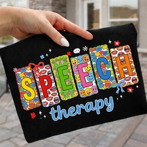 May include: Black t-shirt with the words "SPEECH therapy" in colorful, block letters. Each letter is filled with various images, including lips, hands, and smiley faces. The design is accented with small stars and bows.