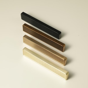 May include: Four rectangular cabinet pulls in black, bronze, brass, and gold. Each pull has a textured, grooved surface and a solid, rounded end. The pulls are arranged diagonally against a neutral background, showcasing their varying metallic finishes.