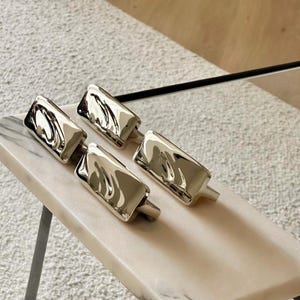 May include: A set of four rectangular cabinet pulls with a polished silver-tone finish. Each pull features a distinct, rippled, and undulating surface texture that creates a reflective, liquid-like appearance across the metallic material.