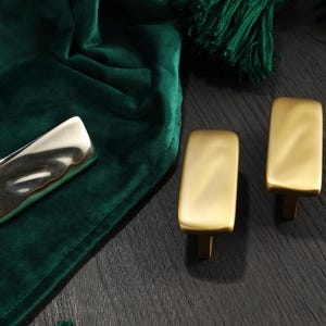 Könnte beinhalten: Eine Sammlung goldfarbener Schrankbeschläge, darunter zwei rechteckige Griffe und ein gebogener Griff. Die Beschläge werden auf einer dunklen Holzoberfläche mit grünem Samtstoff und Quasten im Hintergrund präsentiert.