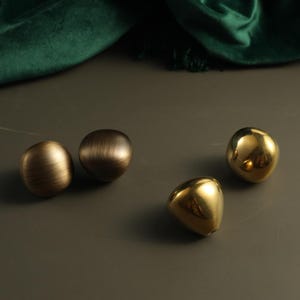 Könnte beinhalten: Vier abgerundete Schrankknöpfe mit unregelmäßigen, organischen Formen. Zwei Knöpfe haben ein gebürstetes, mattes Gold-Finish mit sichtbarer horizontaler Textur, während die anderen beiden eine glatte, polierte, reflektierende Gold-Oberfläche mit Hochglanz aufweisen.