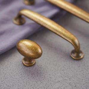 May include: Close-up of antique brass cabinet hardware, including a knob and two pull handles. The hardware has a distressed finish, with visible variations in color. The knob is oval-shaped, and the pulls have a curved design.