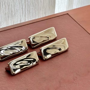 May include: Three rectangular cabinet pulls with a polished silver-tone finish. Each handle features a smooth, undulating, and reflective surface with soft, organic curves that create a distorted mirror effect across the metal face.