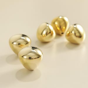May include: A collection of five golden, teardrop-shaped beads. The beads have a smooth, reflective surface, and are arranged on a light-colored background. The beads are likely for jewelry making or other craft projects.