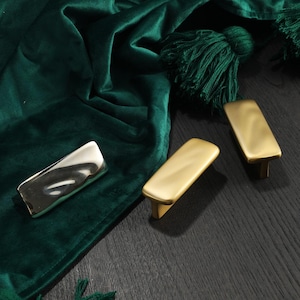Puede incluir: Tres tiradores de armario de diseño moderno. Uno es plateado y dos son dorados. Los tiradores son rectangulares con una parte superior curvada y una base rectangular pequeña. Se muestran sobre una superficie de madera oscura y un paño de terciopelo verde.