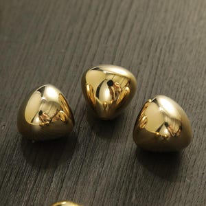 Puede incluir: Tres objetos decorativos dorados brillantes en forma de lágrima. Las superficies lisas y reflectantes crean un efecto de espejo, reflejando el entorno. Los objetos están dispuestos sobre una superficie oscura y texturizada.