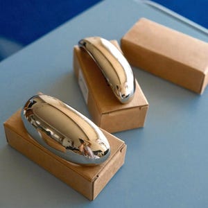 Puede incluir: Tres manijas de puerta cromadas, cada una con forma ovalada y alargada, se presentan sobre pequeñas cajas de cartón marrón. Las manijas pulidas reflejan el entorno. Los artículos están dispuestos sobre una superficie azul claro.