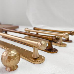 May include: A collection of decorative cabinet pulls in various metallic finishes, including gold and bronze. The pulls feature different knob and handle designs, with textured and smooth surfaces. The hardware is arranged on a white surface, showcasing the variety of styles.