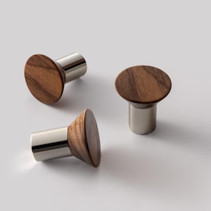 May include: Three wooden cabinet knobs with a circular, dark brown wood top and a silver metal base. The knobs are arranged on a white surface, showcasing their modern design and contrasting materials.