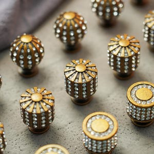 May include: An assortment of antique gold cabinet knobs with crystal accents. The knobs have various shapes, including round and cylindrical, and are arranged on a gray surface. The knobs are likely for furniture or decorative purposes.