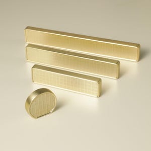 May include: A set of four brass cabinet hardware pieces. The set includes three rectangular bar pulls of varying lengths and a round knob. All pieces have a textured, grid-like pattern on their surfaces.