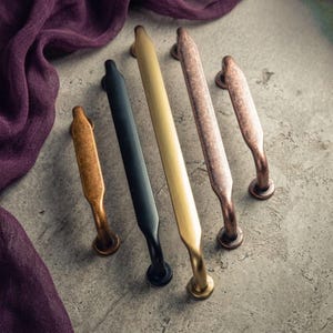 May include: A collection of five metal cabinet pulls in various finishes, including matte black, brushed gold, antique copper, and weathered bronze. Each handle features a central bar with tapered ends and two circular mounting points for attachment.