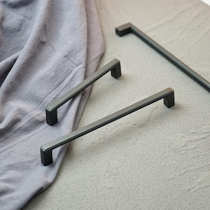 May include: A collection of matte black cabinet pulls in various sizes. The pulls have a modern, minimalist design with a rectangular bar and a square handle. They are arranged on a light gray surface with a gray fabric in the background.