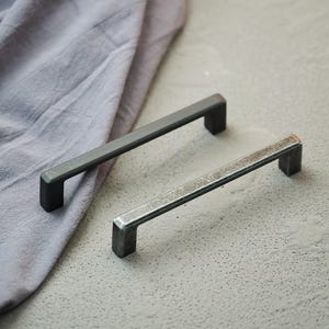 Pode incluir: Duas puxadores de metal para armários. Um é cinza escuro, o outro tem uma aparência envelhecida e desgastada. Ambos têm formato de barra retangular com extremidades quadradas. Estão sobre uma superfície cinza claro com um pano roxo.