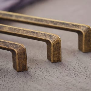May include: Close-up of three antique brass cabinet pulls. The pulls have a rectangular design with rounded corners and a distressed finish. The hardware is a warm, aged brass colour, perfect for adding a vintage touch to furniture or cabinetry.