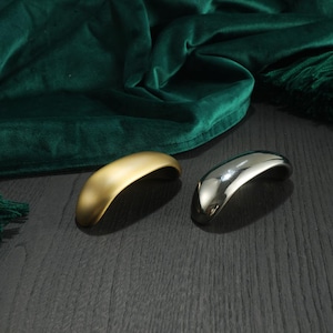 May include: Two curved cabinet pulls, one gold and one silver, rest on a dark wood surface. The gold pull has a matte finish, while the silver one is highly reflective. A dark green velvet fabric is in the background.