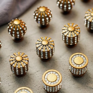 Peut inclure: Gros plan de plusieurs boutons de meubles décoratifs. Les boutons sont dorés avec des accents de cristaux. Certains sont sphériques, d'autres cylindriques ou avec un motif de soleil. Les cristaux sont clairs et réfléchissent la lumière.