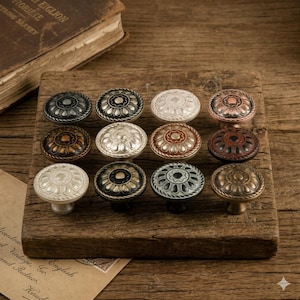 May include: A collection of twelve decorative cabinet knobs in various metallic finishes, including bronze, silver, and copper. Each knob features a floral design and is displayed on a wooden surface, showcasing a vintage aesthetic.