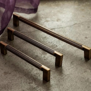 May include: Three metal drawer pulls in varying lengths, each featuring a dark bronze finish with a ribbed, cylindrical center bar and smooth, rectangular mounting posts at both ends, arranged diagonally from shortest to longest.