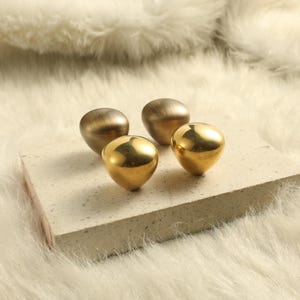May include: Four teardrop-shaped cabinet knobs arranged in a cluster. Two knobs feature a polished, reflective gold finish, while the other two knobs have a brushed, matte metallic finish with visible horizontal graining and a muted tone.
