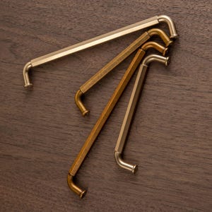 May include: Four metal cabinet pulls in shades of gold and bronze. Each pull has a straight, hexagonal bar that curves at the ends. The pulls are arranged diagonally on a dark wood surface.