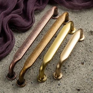 May include: Four metal cabinet pulls arranged in a row, featuring varying finishes including a dark copper, a distressed antique brass, a bright brushed gold, and a matte champagne gold, each with a curved handle and two mounting posts.