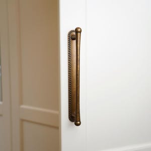 May include: A bronze-colored door pull with a decorative beaded edge and rounded ends. The handle is mounted on a white door, showcasing its vertical design and antique aesthetic.