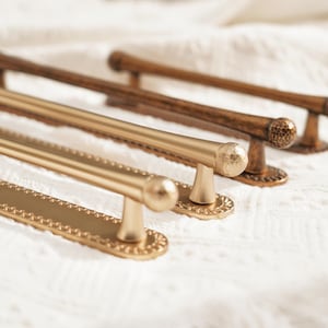 May include: A selection of cabinet pulls in gold and brown finishes. The gold pulls have a decorative edge and a rounded handle. The brown pulls have a textured handle and a rounded end. The pulls are designed for furniture.