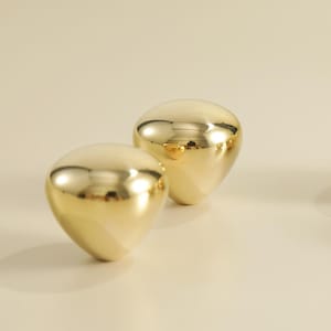 May include: Two golden, teardrop-shaped decorative objects with a polished, reflective surface. The objects are positioned on a light-colored surface, reflecting the surrounding light. The smooth, rounded forms suggest a modern, minimalist design.