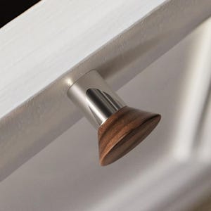 May include: A modern cabinet knob with a cylindrical brushed metal body and a conical, dark wood handle. The knob is mounted on a white surface, creating a contrast between the materials and colors.