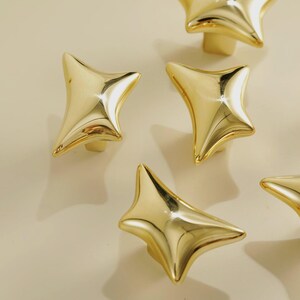 May include: Close-up of several gold-colored star-shaped cabinet knobs. The knobs have a shiny, reflective surface and a four-point star design. The knobs are likely made of metal and are designed for furniture or cabinetry.