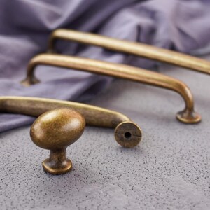 Op de afbeelding: Een set antiek messing deurbeslag bestaande uit twee lange, gebogen kastgrepen en één ovale kastknop. Elk stuk heeft een verweerde, metalen afwerking met een warme, goudbruine tint en een glad, afgerond profiel voor meubels.