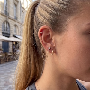 May include: A gold chain earring featuring two delicate chains connecting a small stud at the upper ear cartilage to a four-stone cluster stud on the earlobe, accompanied by a separate small gold hoop earring worn on the same earlobe.