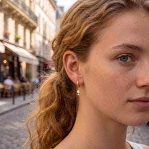 May include: A gold hoop earring featuring a small, round, iridescent stone charm suspended from the bottom. Below the stone is a tiny, four-pointed star-shaped metal accent. The earring is worn in the earlobe, showcasing a polished gold finish.