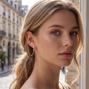 May include: A gold-toned ear cuff connected by a delicate chain to a stud earring. The ear cuff features a row of small clear stones, while the stud earring displays three vertically aligned clear stones set in a gold-toned metal base.