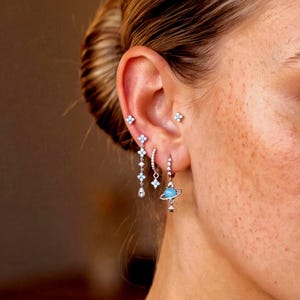 May include: A set of silver-toned earrings featuring a dangling chain with four-petaled flower charms, a small hoop with a star charm, and a hoop earring with a blue planet charm surrounded by a ring, plus two small flower stud earrings.